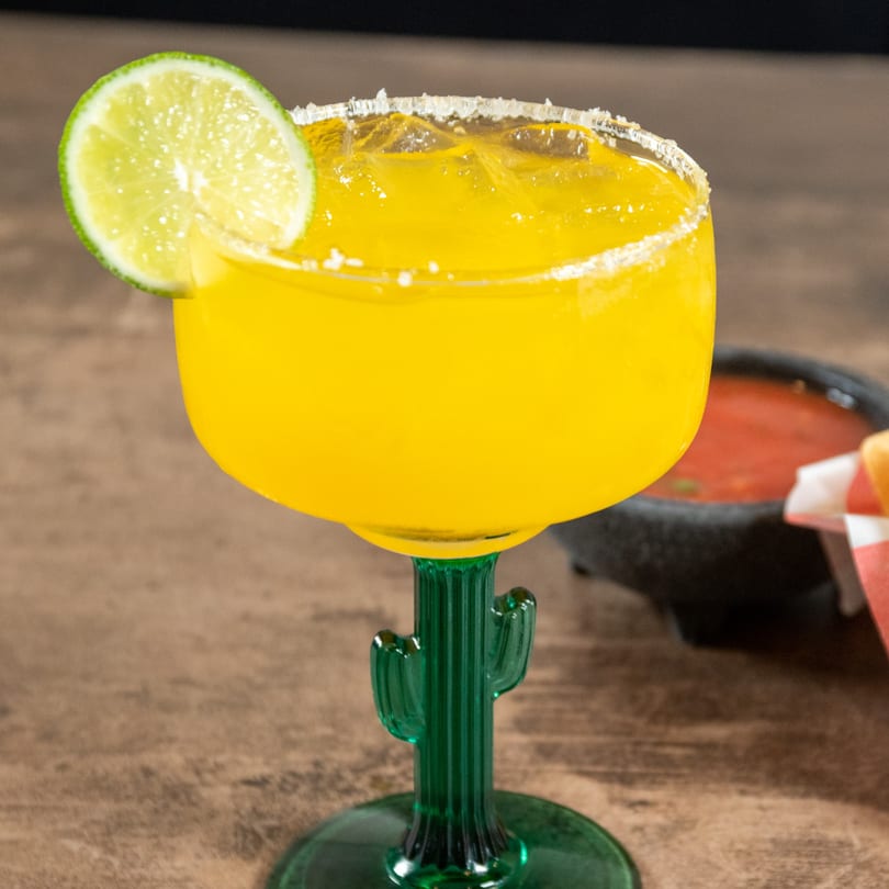 A vibrant yellow margarita with a salted rim and lime wedge, served in a cactus-stemmed glass.