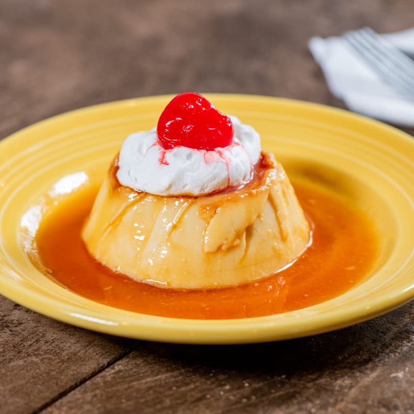 Flan dessert with whipped cream and a cherry on top, served in a yellow dish.